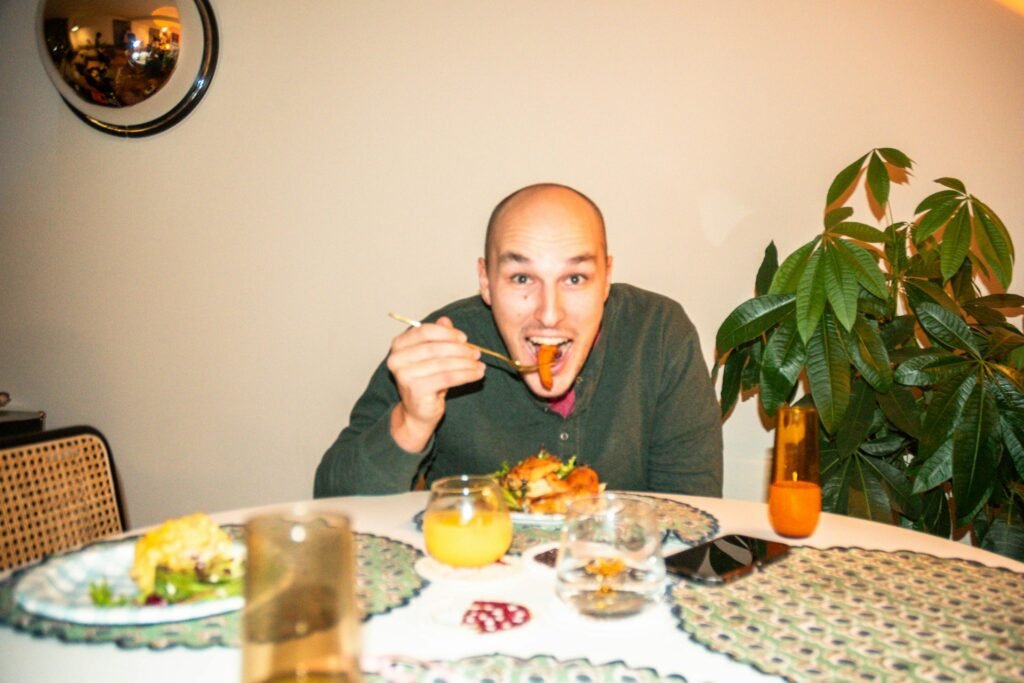 A smiling man sitting at a dining table, enthusiastically eating with chopsticks. The table is set with plates, glasses, and a drink, while a green potted plant decorates the background, adding a cozy and inviting atmosphere.